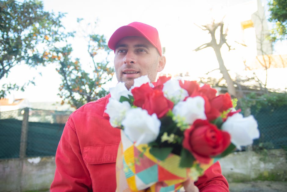 A male florist dressed in a red cap and matching red uniform holding a colorful bouquet of fresh flowers, including red and white roses and other blooms, wrapped in vibrant patterned paper. The arrang
