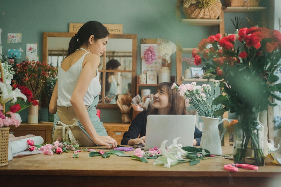 A young female florist with dark hair tied back, wearing a white sleeveless top and beige apron, is standing at a wooden work table inside a flower shop displaying a variety of floral arrangements. Sh