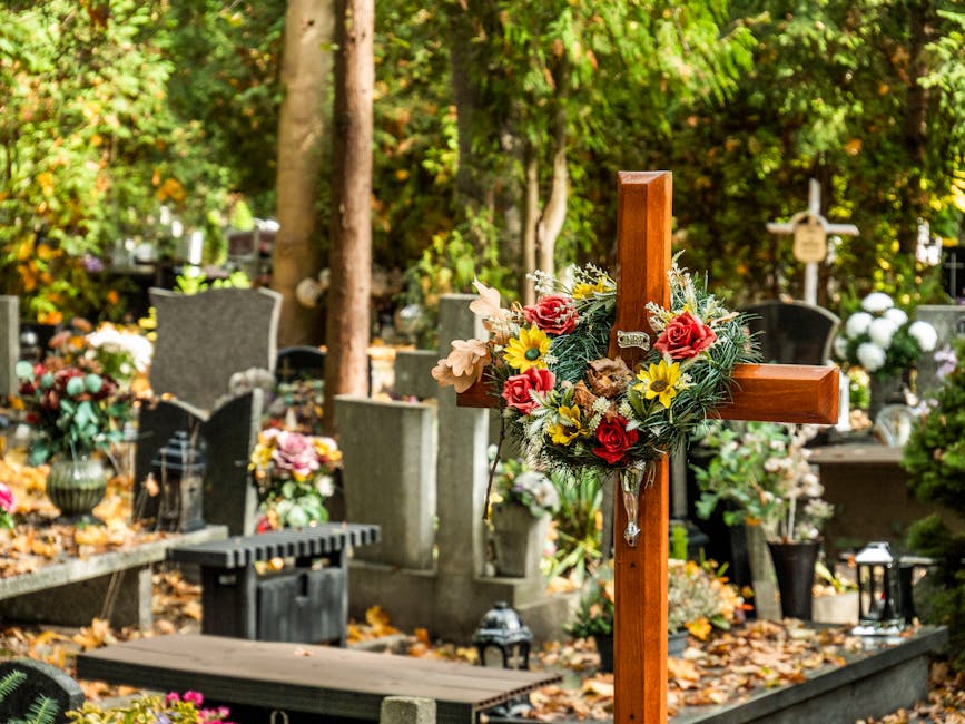A wooden cross adorned with a floral arrangement featuring red roses, yellow daisies, pale pink flowers, and greenery, positioned among gravestones and memorial plaques in a cemetery. Surrounding the 