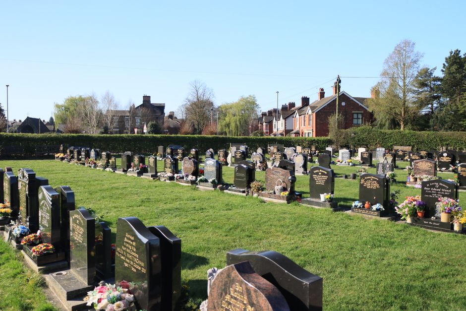 A peaceful cemetery scene featuring rows of dark granite headstones, each decorated with fresh floral arrangements including colorful bouquets of roses, daisies, and lilies. The flowers display a rang