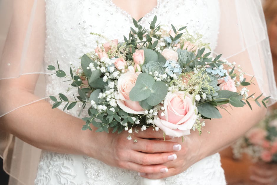 A bride holding a floral bouquet featuring pale pink roses, white baby's breath, and eucalyptus leaves, arranged in a natural, loosely styled composition. The bouquet is wrapped in soft, light-colored